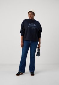 Granatowy sweatshirt z logo, luźny krój; zestawiony z dzwonowymi niebieskimi dżinsami. Dodatki to mała torebka i jasno-beżowa czapka. Czarne buty.
