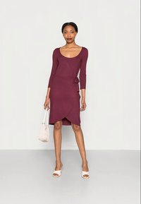 Vestido envolvente bordeaux com mangas longas, um detalhe atado na cintura e uma bainha até ao joelho. Apresenta um decote arredondado e uma textura suave.