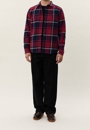 Mann trägt ein rot-navy kariertes Hemd mit Knöpfen, schwarze Hose und braune Schuhe, steht vor einem einfarbigen hellen Hintergrund.