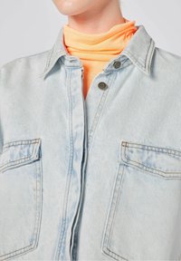 Camicia di denim azzurro chiaro con due taschini sul petto, bottoni a pressione e un morbido dolcevita arancione sottostante; design casual con una vestibilità rilassata.