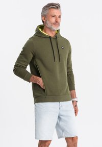 Sudadera verde oliva con un bolsillo canguro delantero, cordones negros y acentos en neón amarillo. Combinada con pantalones cortos de mezclilla claros.