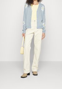 Žena v světle modrém kardiganu s bílými sedmikráskami, žlutém tričku, krémových kalhotách, béžových botách a nese malou žlutou kabelku.