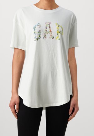 Biała koszulka z krótkim rękawem z napisem "GAP" utworzonym z wielokolorowych kwiatowych liter, noszona przez osobę stojącą na jednolitym tle.