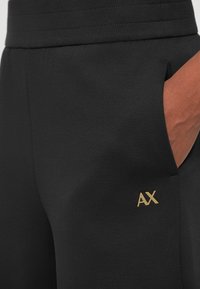 Calças de moletão pretas com um cós canelado, apresentando um bolso de detalhe castanho. Detalhe de logótipo em dourado na lateral, ressaltando a textura suave do tecido.