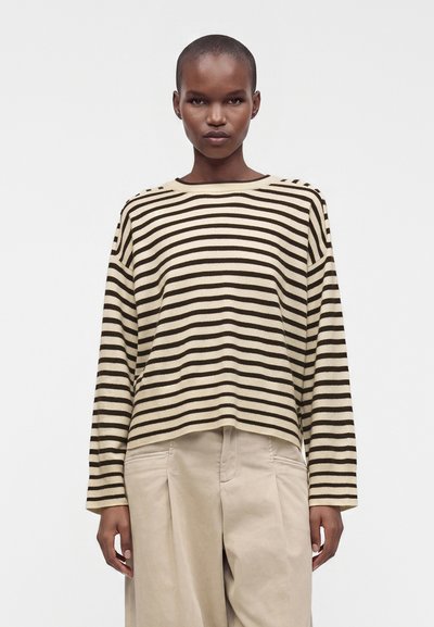 Personne portant un haut à manches longues beige et noir à rayures horizontales et un pantalon beige à jambes larges, debout devant un fond blanc uni.