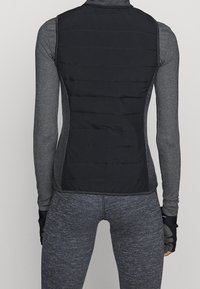 Czarna kamizelka puchowa z teksturą podszewki, noszona na szarym longsleeve. Posiada dopasowany krój i płaskie szwy dla str streamlined appearance.