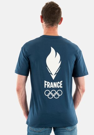 T-shirt bleu marine à manches courtes avec un design de flamme olympique blanche, le texte "FRANCE", et cinq anneaux entrelacés au dos.
