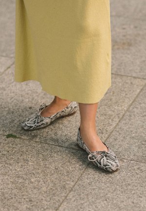 Pieds portant des ballerines à motif peau de serpent avec des nœuds, debout sur un pavé en pierre, partiellement recouverts par une jupe midi jaune.