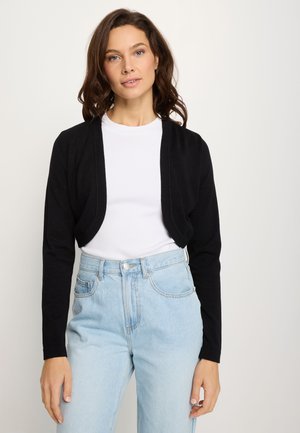 Woman with wavy brown hair wears a black cropped cardigan over a white top and light blue high-waisted jeans, standing against a plain background.