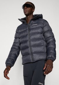 Muškarac u crnoj puffer jakni i hlačama s Berghaus logotipom, s crnim naočalama za sunce, stoji nasuprot jednostavne svijetle pozadine.