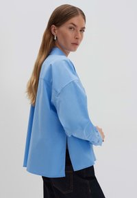 Camicia oversize azzurro chiaro in cotone con colletto, fessure laterali e polsini con bottoni, abbinata a jeans scuri.