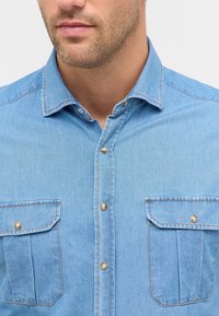 Lichtblauw denim overhemd met een button-down kraag, twee voorzakken en contrasterende stiksels. Voorzien van bronzen knopen voor sluiting.