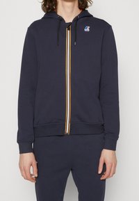 Námořnicky modrá mikina s kapucí vyrobená z bavlněné směsi, vybavená předním zipem se žluto-bílými pruhy, žebrovanými manžetami a logem na hrudi.