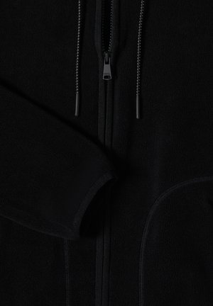 Close-up van een zwart fleecejack met rits aan de voorkant, trekkoorden en zichtbaar textuurstof op de mouw en rand van de zak.