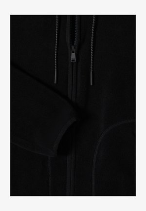 Close-up van een zwart fleecejack met rits aan de voorkant, trekkoorden en zichtbaar textuurstof op de mouw en rand van de zak.