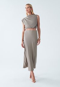 Slank, todelt outfit i lys taupe. Har en cropped, draperet top og en lang, flydende nederdel med en glat tekstur. Sandaler i gyldne nuancer.