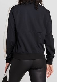 Person iført en sort jakke med hvide ærmer og sorte leggings med et hvidt stjernedesign, vist fra ryggen.