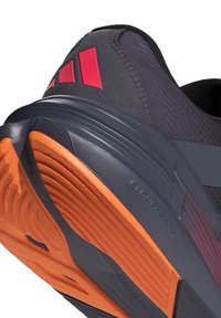 Nahaufnahme eines dunkelgrauen Sportschuhs mit oranger Sohle und rotem Logo, mit strukturiertem Grip und gepolstertem Fersendesign.