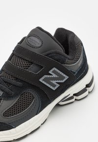 Chaussure de sport noire en matériaux mesh et daim, avec des brides Velcro, un accent de logo gris et une semelle blanche contrastante.