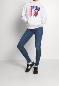 Sudadera blanca con texto gráfico en rojo y azul, vaqueros ajustados azul, y zapatillas negras con rayas blancas. Tela suave, estilo casual.