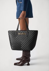Crna torba od kviltane tkanine s uzorkom chevron, zlatnim logom i elegantnim crnim ručkama. Uparena s smeškanim kožnim gležnjačama.