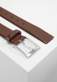 Bruine leren riem met een gladde textuur, uitgerust met een zilveren rechthoekige gesp met een reliëflogo en gaten van een inch breed voor maataanpassing.