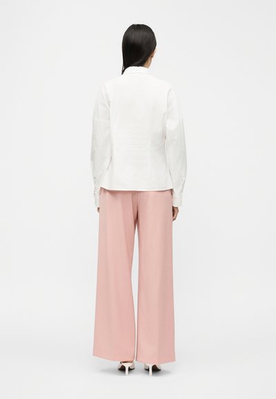 Άτομο με μακριά σκουρόχρωμα μαλλιά που φοράει λευκό πουκάμισο με μακριά μανίκια, κάτω μέρος με φαρδιά ροζ παντελόνια και λευκά παπούτσια με ψηλό τακούνι, γυρισμένο πλάτη.