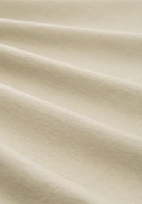 Beige színű pamut anyag, amely puha textúrával rendelkezik, finom hullámokkal és ráncokkal, hangsúlyozva könnyedségét és sima felületét.