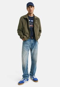 Olive green bomber jacket over a navy graphic T-shirt, light blue jeans, and blue athletic sneakers with white accents. Blue cap completes the look.