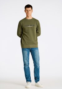 Olivgrön sweatshirt med rund halsringning, med "COPENHAGEN LINDBERGH" i marinblått tryck. Kombinerad med ljusblå jeans och vita sneakers.