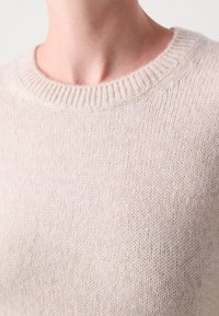 Gros plan d'une personne portant un pull beige en maille douce avec un col rond côtelé.