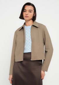 Γυναίκα με κοντά σκούρα μαλλιά που φορεί ανοιχτό γαλάζιο πλεκτό πουλόβερ, κοντό μπεζ σακάκι και σκουρό καφέ σατέν φούστα σε απλό λευκό φόντο.