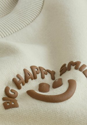 Cremfarbenes Stoffmaterial mit geripptem Kragen, geprägt mit braunem Text "BIG HAPPY SMILE" und einem lachenden Smiley-Design. Weiche Textur.