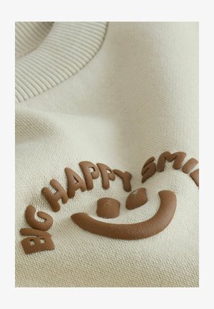 Kreminės spalvos audinys su gofruota apykakle, ant kurios iškilūs rudi žodžiai „BIG HAPPY SMILE“ ir šypsenėlės dizainas. Minkšta tekstūra.