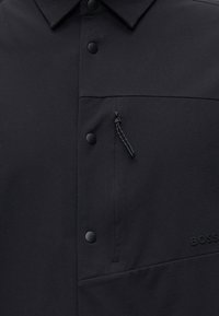 Gros plan d’une veste noire avec des boutons-pression noirs mats, une poche poitrine zippée et un logo BOSS embossé sur le côté droit.