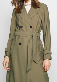 Kobieta ubrana w podwójnie zapinaną płaszcz w oliwkowym kolorze z czarnymi guzikami i pasującym paskiem przewiązanym w talii.