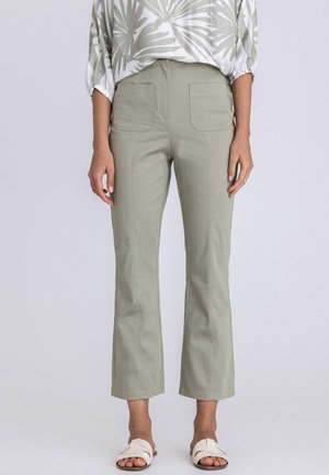 Vrouw draagt lichtgroene cropped broek met voorzakken, witte instapsandalen en een witte blouse met bladmotief.