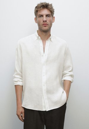 Blond man with light stubble wearing an off-white button-up shirt with rolled sleeves and black pants, standing against a plain light background.