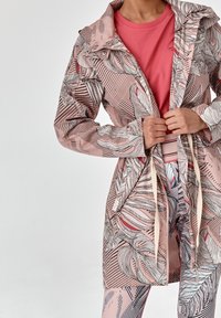 Jasnoróżowy płaszcz przeciwdeszczowy z czarno-białymi wzorami liści, regulowany ściągacz w talii, detale z guzikami oraz warstwowa tekstura materiału.