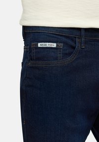 Dark blue denim jeans feature a patch pocket with white label detailing. The fabric has a soft, textured finish and contrasting stitch accents.
