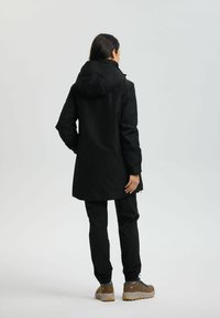 Schwarze wasserdichte Jacke mit Kapuze, langer Schnitt und lockerer Passform, kombiniert mit schwarzen Hosen und braunen Wanderschuhen.