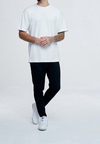 Man som står med benen korsade, iklädd en vit oversized t-shirt, svarta byxor, vita sneakers, med en tatuerad höger underarm synlig.