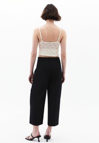 Krämfärgad spetscroptop med justerbara axelband, kombinerad med högmidjade svarta vida byxor och svarta strapponklackar, sedd bakifrån.