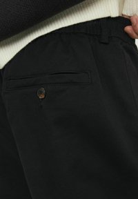 Pantaloni neri realizzati in tessuto testurizzato, dotati di una fascia elastica in vita e una tasca posteriore con chiusura a bottone. Design semplice e funzionale.