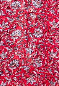 Camicia rossa con motivi floreali blu e bianchi intricati, dotata di bottoni rossi lungo il davanti e un tessuto morbido e testurizzato.