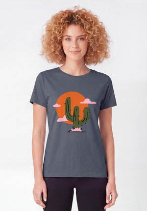 Femme aux cheveux bouclés portant un t-shirt gris avec un cactus vert, des nuages roses et un soleil orange en arrière-plan.