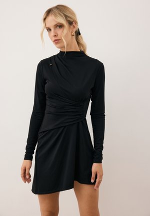 Zwart, lange mouwen jurk met plooien bij de taille, hoge halslijn en een decoratieve gouden accent. Gemaakt van zachte, rekbare stof.