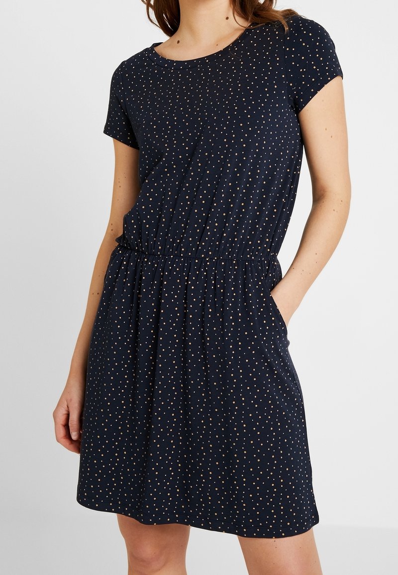 Femme portant une robe bleu marine à manches courtes avec des petits pois blancs et une taille élastique, debout avec une main dans une poche.