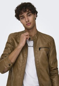 Giubbotto in pelle marrone con un design aderente, dotato di tasca sul petto con zip e colletto a scatto, indossato sopra una T-shirt bianca.