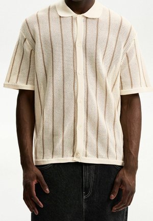 Mand iført en cremefarvet kortærmet strikket skjorte med knapper og lodrette brune striber samt mørke bukser, stående foran en hvid baggrund.